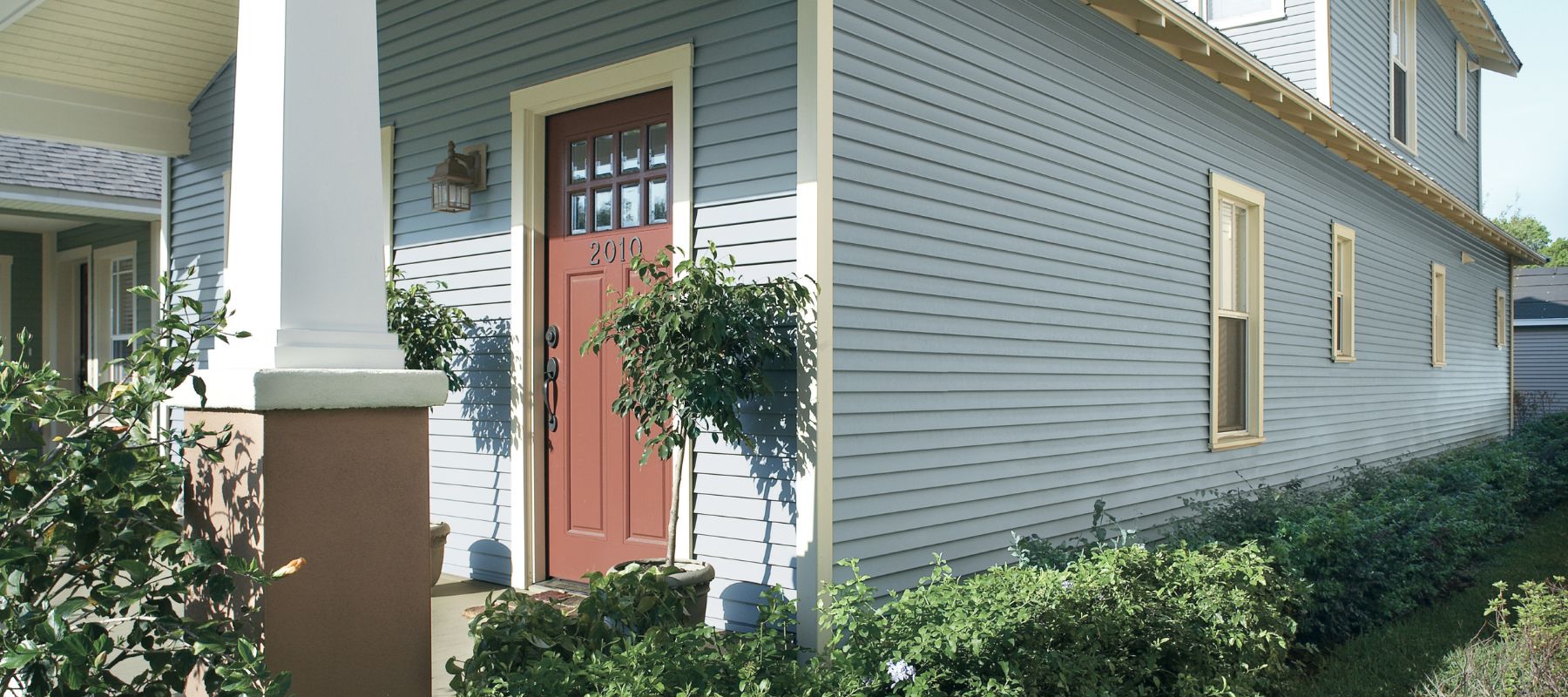 A light blue house with horizontal siding features a red front door with small window panes, white trim, and a welcoming front porch with a large column