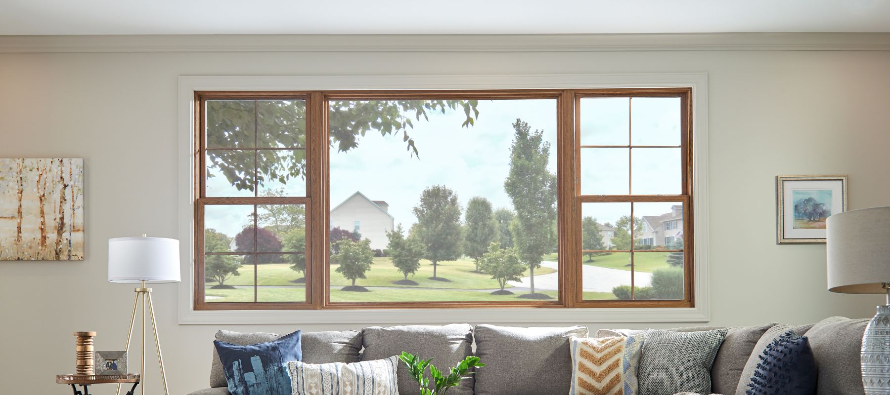 A wide, multi-pane window with wooden frames looks out onto a suburban landscape with trees and houses, viewed from a living room with a sofa and lamps