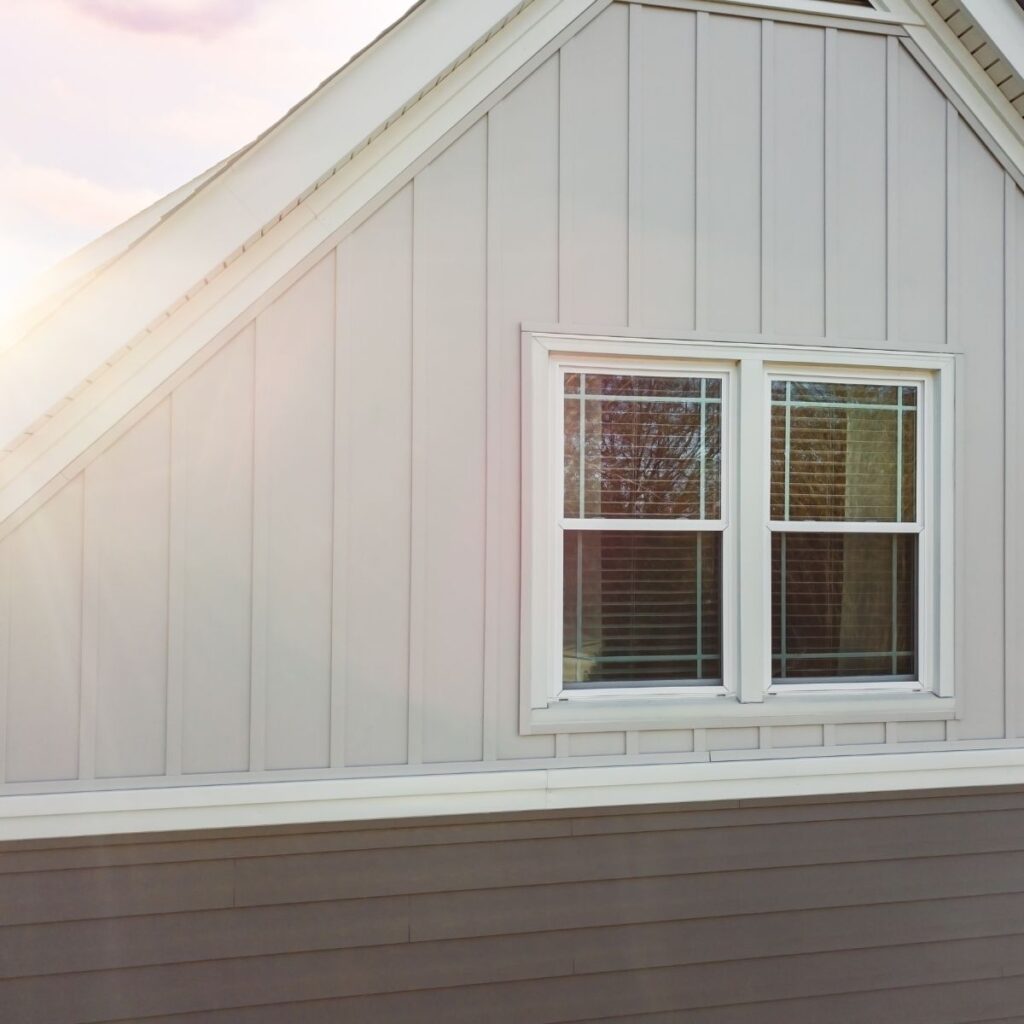 A close-up of a house exterior shows a white-trimmed window with blinds, set into light grey vertical and horizontal siding
