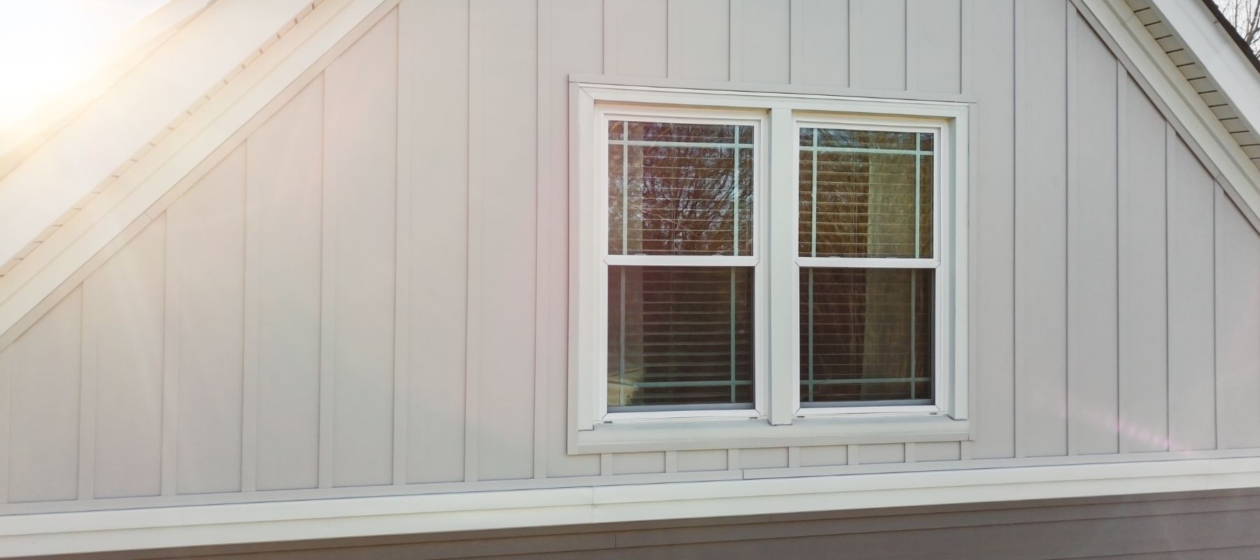 A close-up of a house exterior shows a white-trimmed window with blinds, set into light grey vertical and horizontal siding